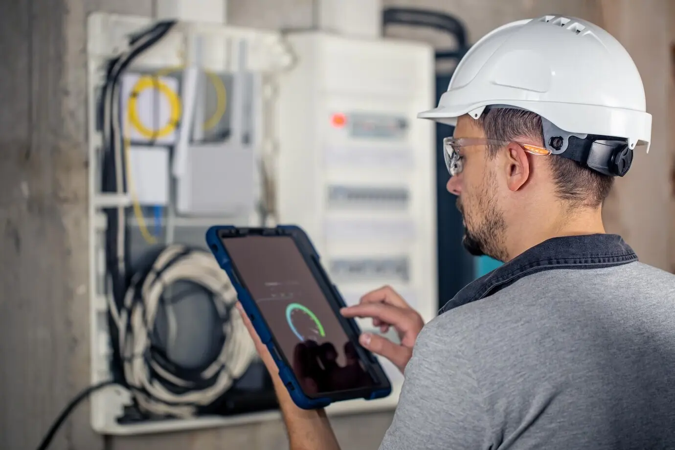 A man, an electrical technician, is working on a switchboard with fuses while using a tablet.