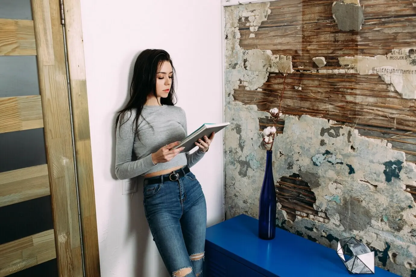 A stylish young woman leans against the wall, reading a journal.